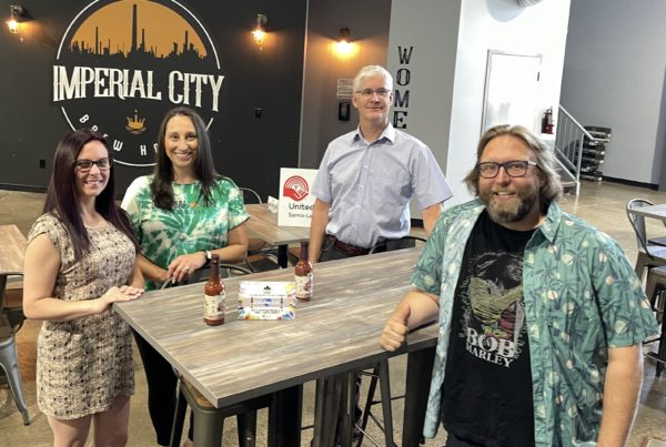 United Way, Imperial City Brew House, and Top Shelf Canada representatives pose infront of the Imperial City logo on the wall behind them, displaying hot sauce bottles