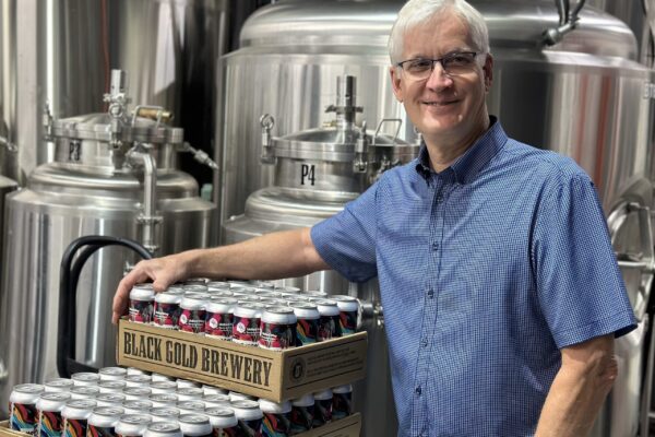 United Way board member Alan Blahey poses with three cases of United Way beer, with large beer kegs in the background